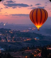 A hot-air balloon drifting over a hillside city at dusk as the street lights come on.