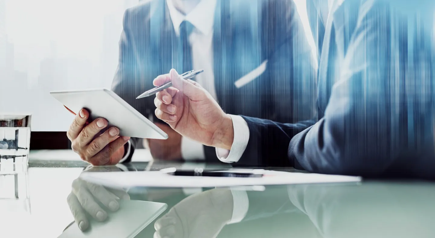 Two colleagues in business dress reviewing figures on a tablet across a glass meeting table.